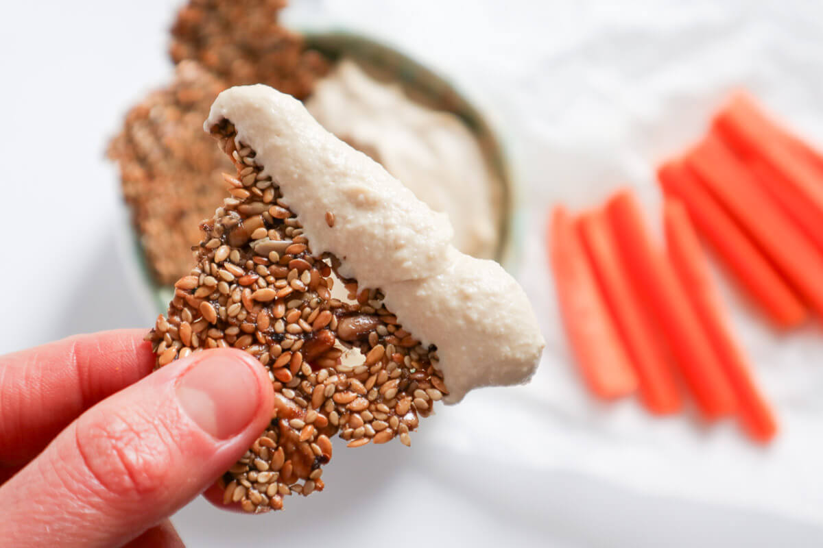 Sunflower sesame seed crackers held by hand and dipped in hummus