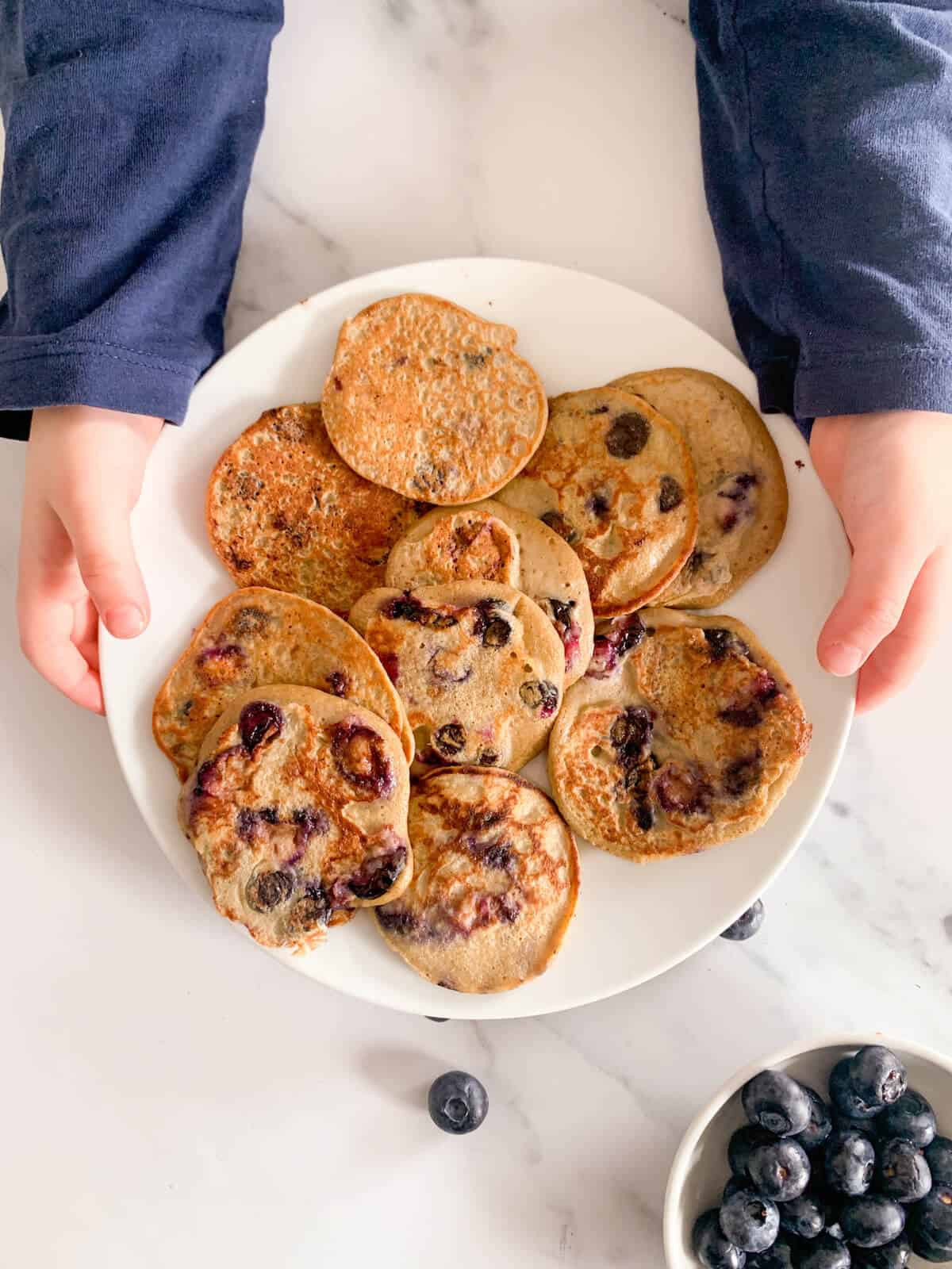 Toddler holding plate with lots of healthy blueberry pancakes