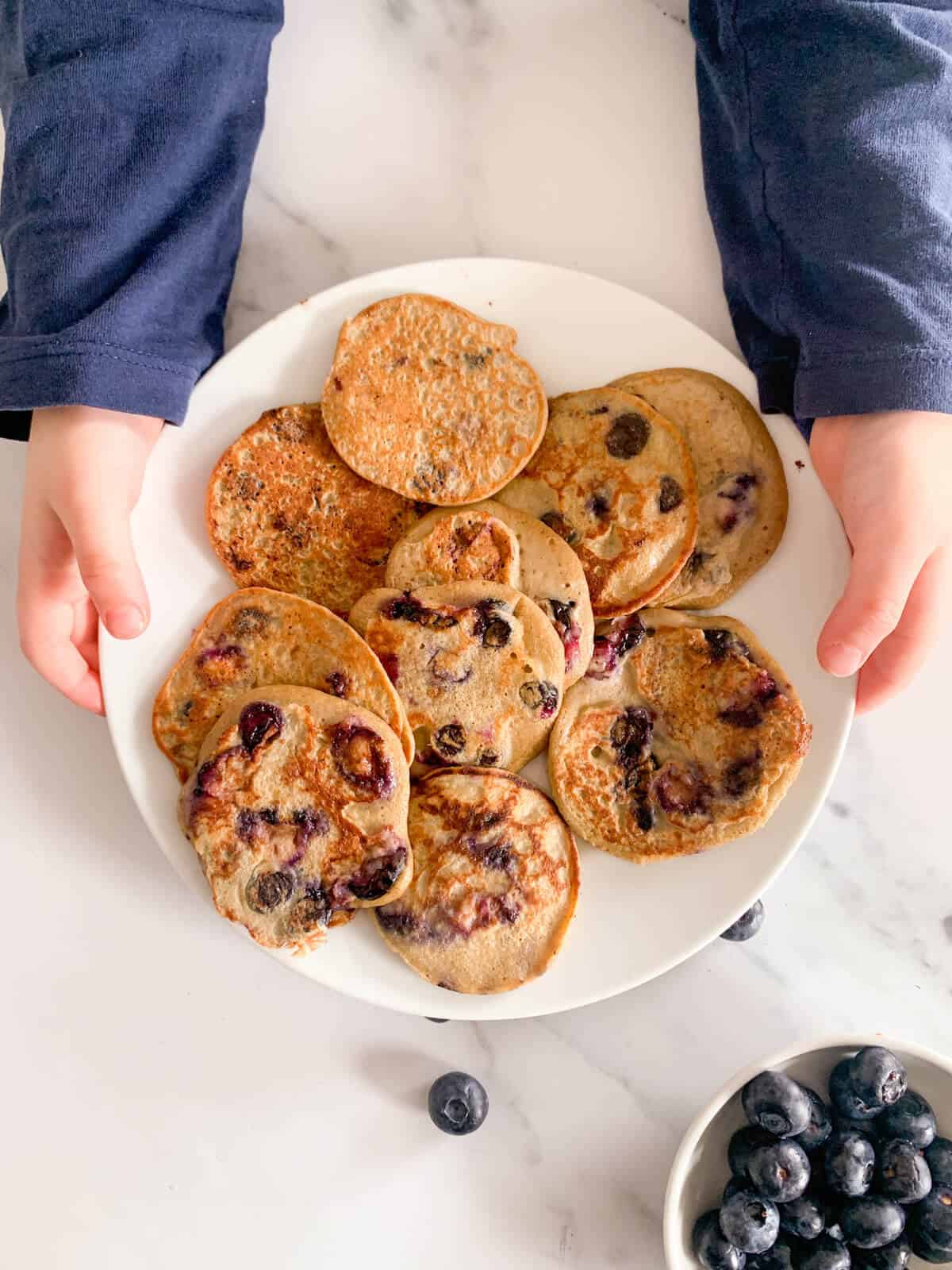 Toddler holding plate with lots of healthy blueberry pancakes 