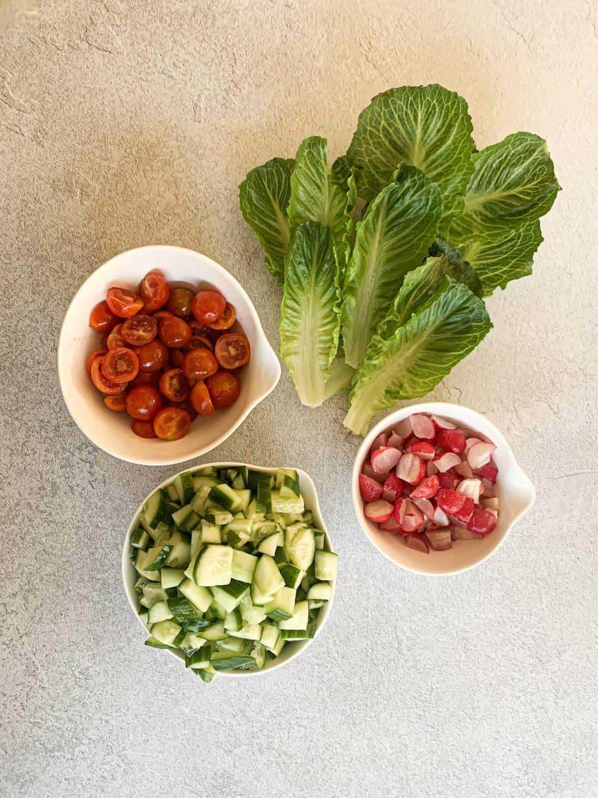 salad ingredients - lettuce, bowl of cherry tomatoes, bowl of chopped up cucumber, bowl of chopped up radishes 