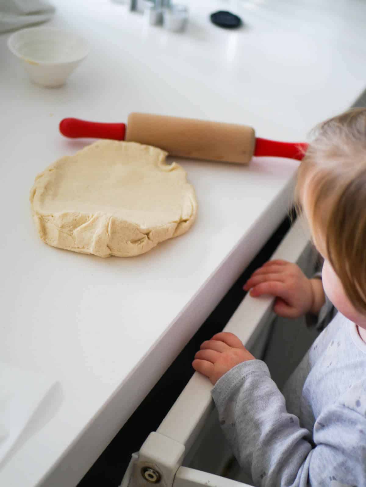 toddler standing in learning tower looking at dough on the counter 