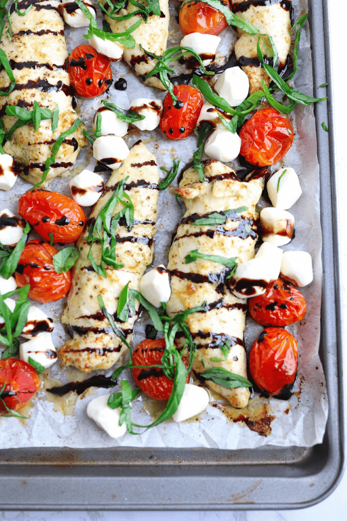 Baked chicken tenders on parchment paper in a pan with mozzarella, cherry tomatoes and basil. 