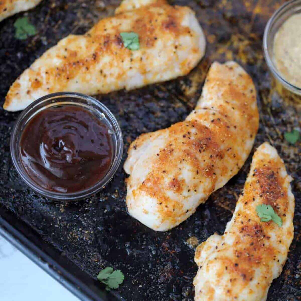 Three chicken tenders on a black plate with 2 sauces on the side.