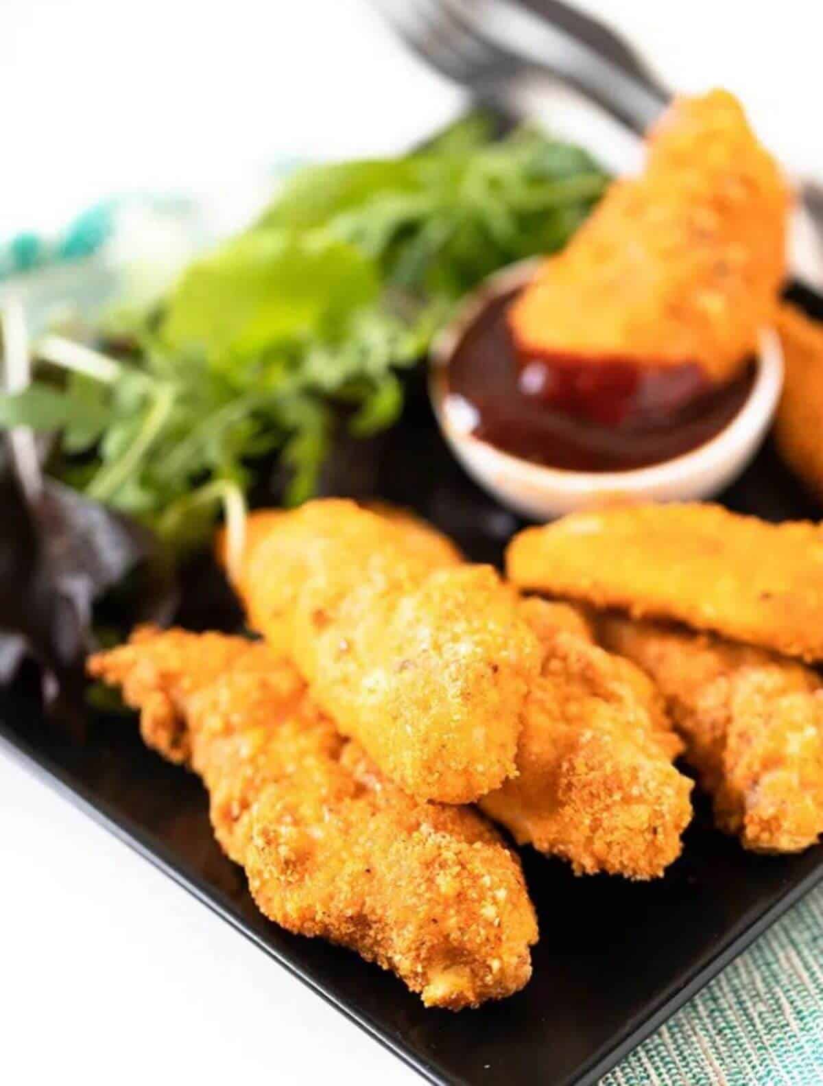 Baked chicken tenders on a black plate with sauce and salad on the side. 