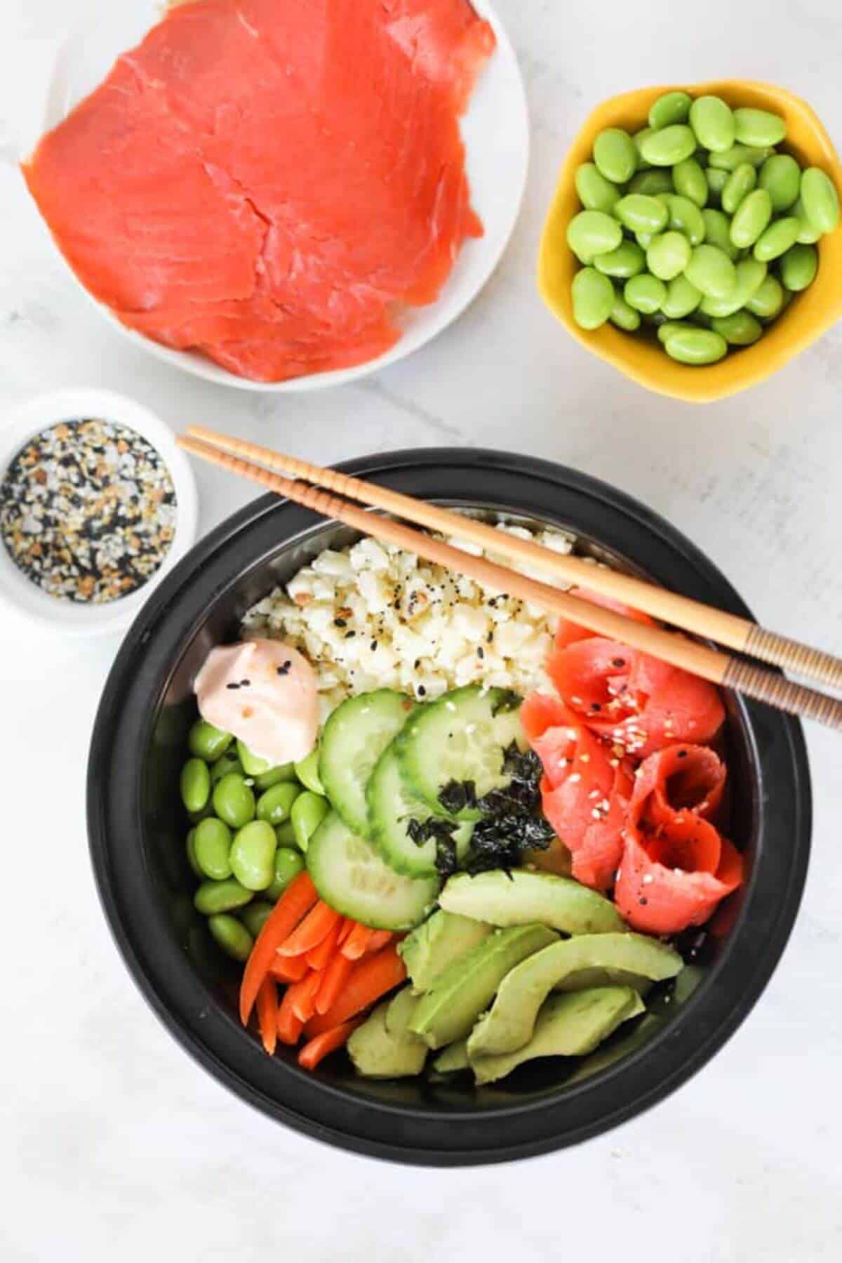 A black bowl with smoked salmon, avocados, carrots, edamame, cucumbers and rice with 2 chopsticks. 