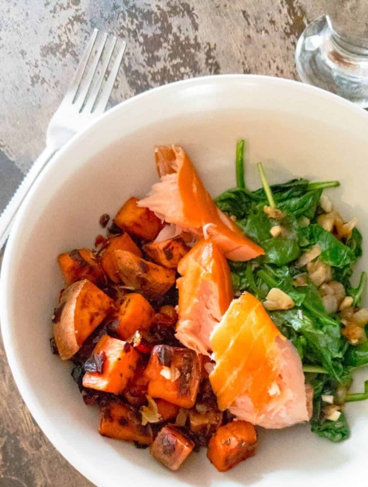 White bowl with smoked salmon, sweet potato and leafy greens. 