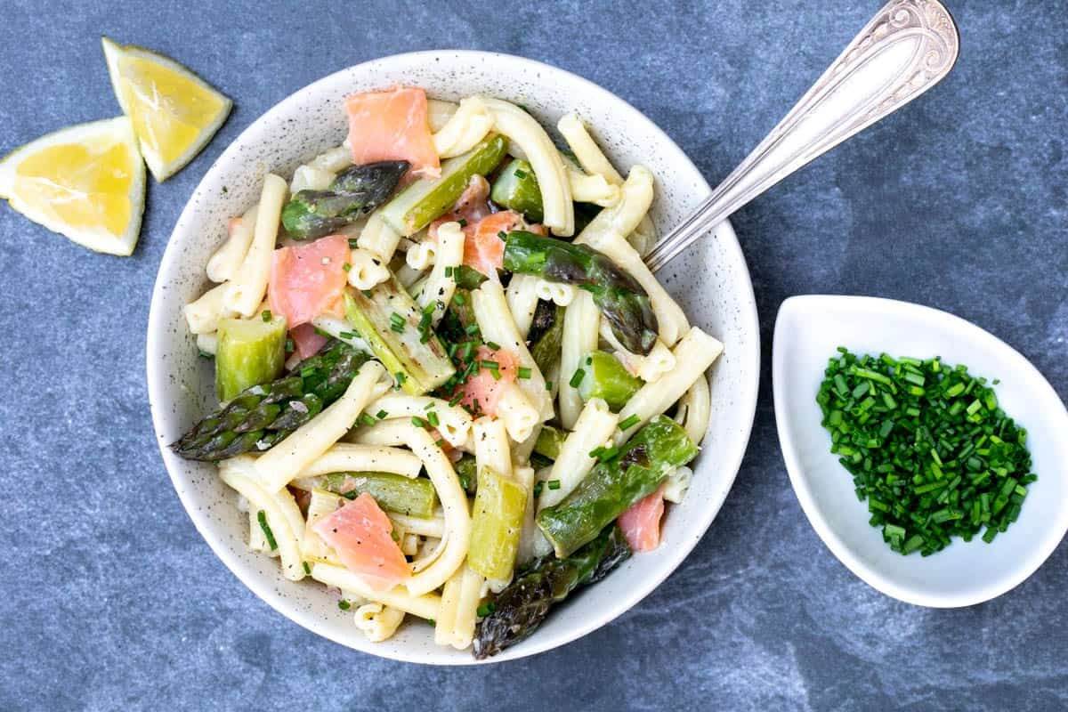 White plate with pasta, asparagus and smoked salmon with some herbs for garnish and lemon slices on the side. 