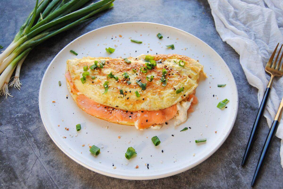 White plate with an omelet filled with smoked salmon and some spring onions on the side. 