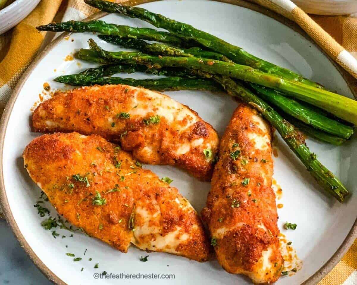 Three baked chicken tenderloins on a plate with asparagus on the side. 