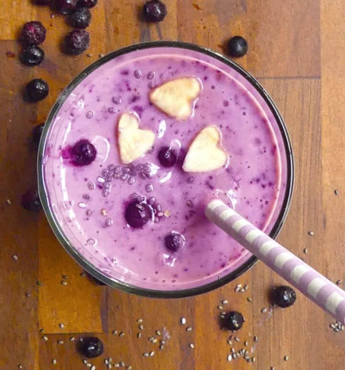 Purple smoothie with chia, blueberries and banana-shaped hearts on top. A purple straw inserted. 