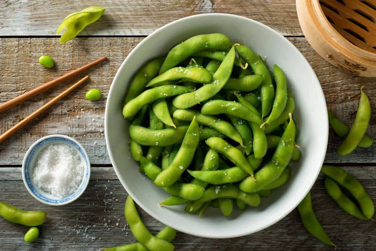 Edamame in a bowl, sprinkled with salt, in a bowl on a table next to a few other edamame, a pair of chopsticks and a bowl of salt