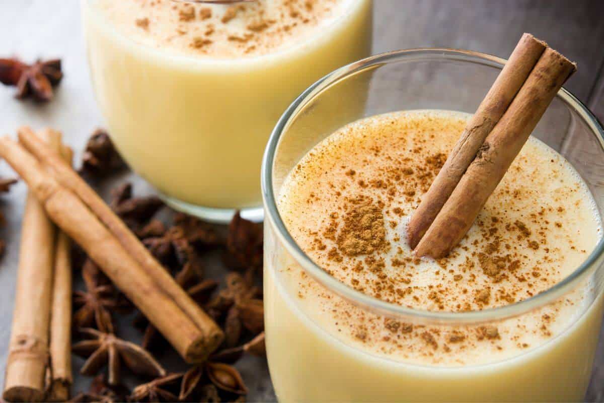 2 glasses of eggnog, with a cinnamon stick sticking out, dusted with ground nutmeg and further cinnamon sticks and star anise on the table next to them