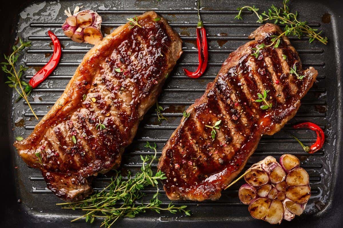 2 eland steaks on a grill surrounded by a few herbs, chillis and roasted garlic