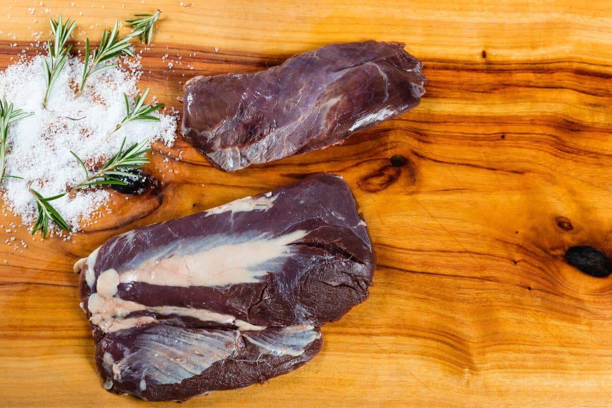 2 emu steaks on a wooden board next to some salt and rosemary