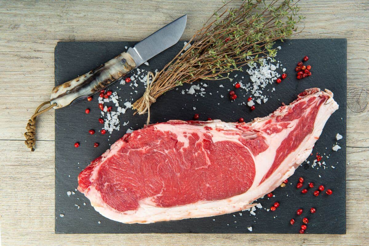 A raw entrecote steak on a black slate board, next to some sea salt, herbs, red berries and a knife