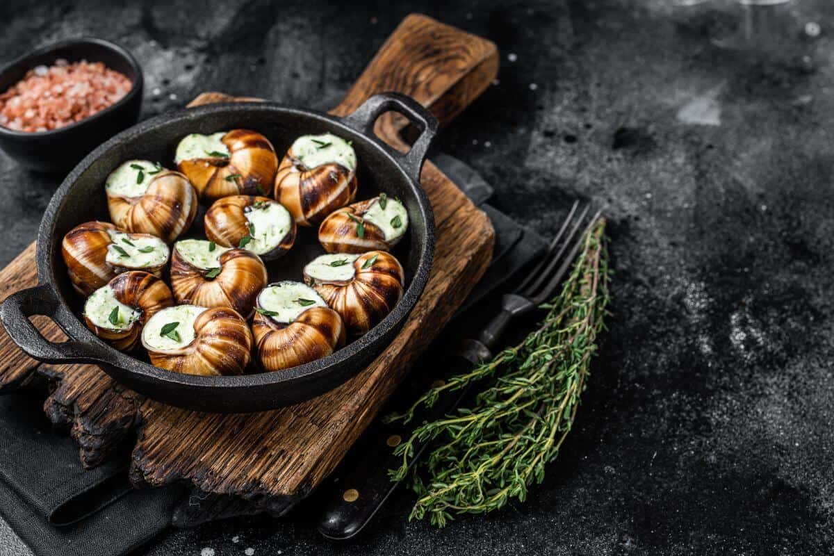 Escargot, covered in garlic butter, ina cast iron dish sitting on a wooden board