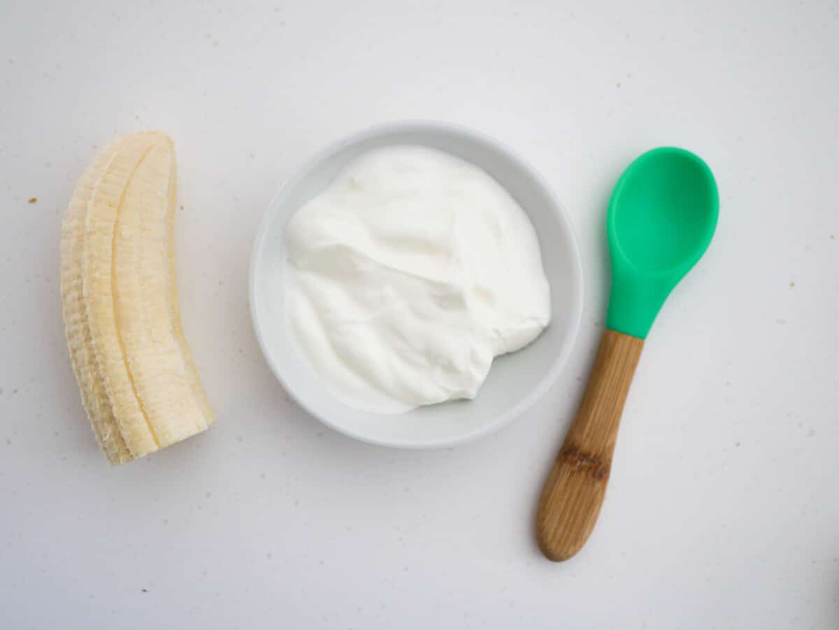 half banana, yogurt in a bowl, baby silicone spoon in green 