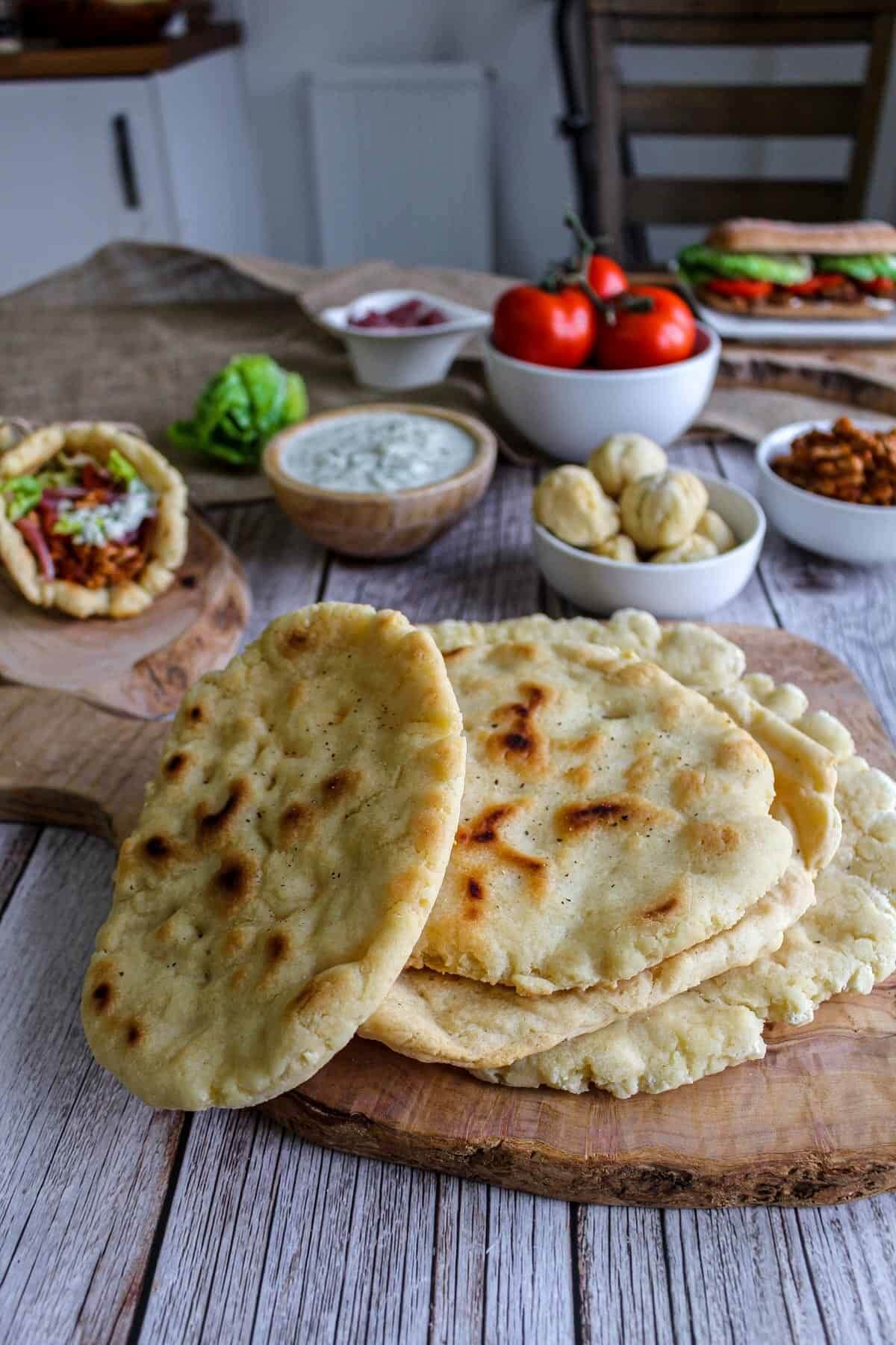 Four pieces of round flatbread on a wooden plate with finger food and dips in the background. 