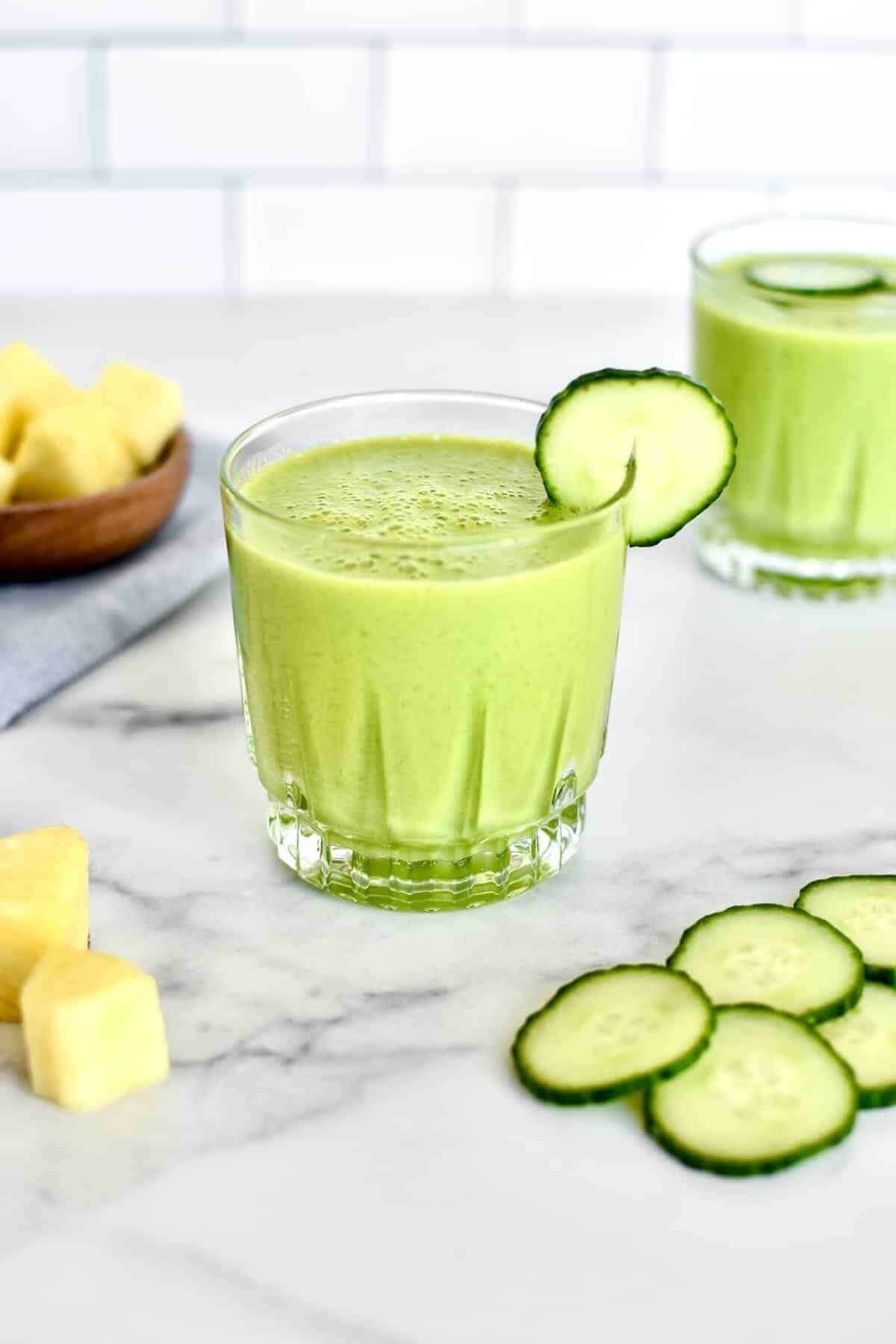 Glass with a green smoothie and cut up cucumbers and mangoes on the side. 