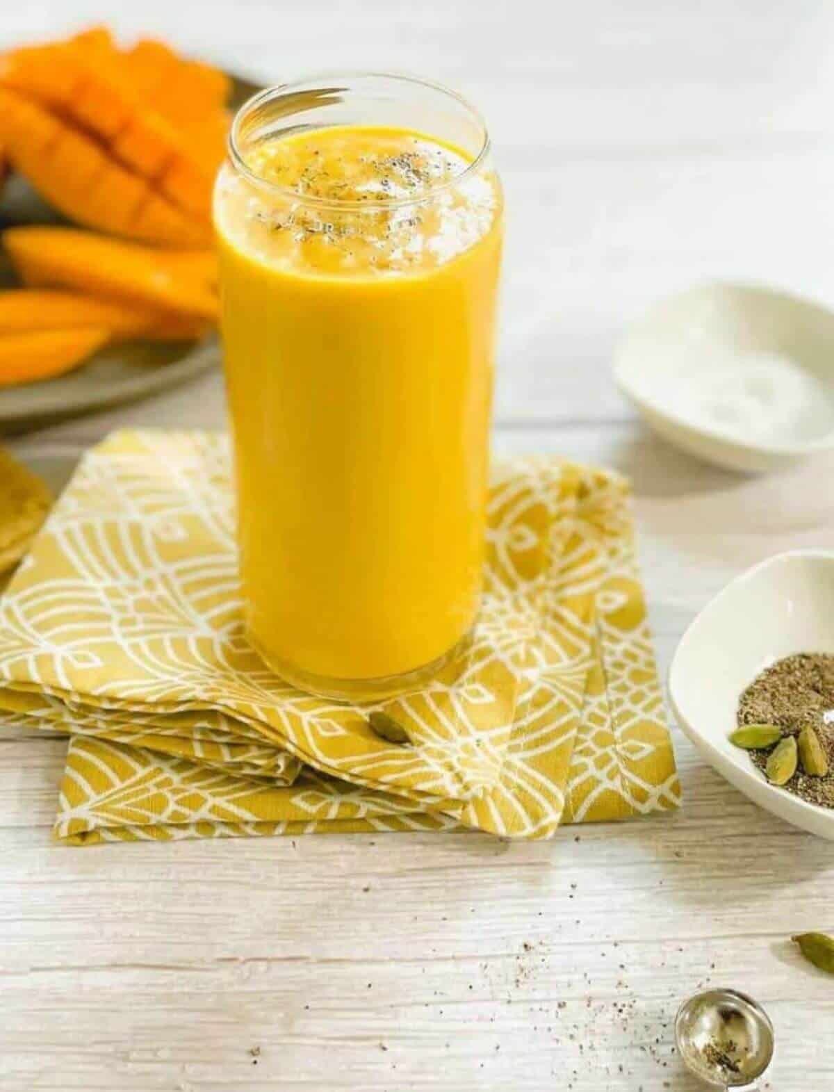 Glass with a yellow smoothie on a yellow cloth and mangoes in the background. 