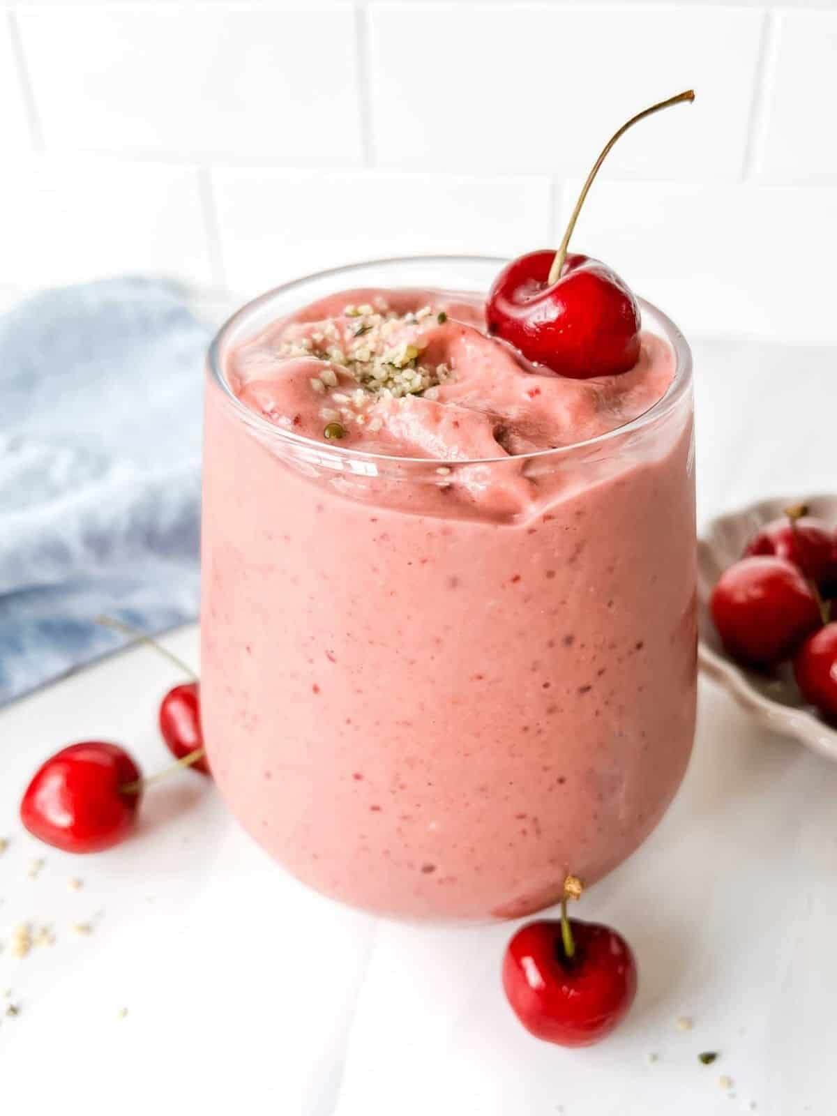 Glass with a pink smoothies and one cherry on top and three on the side. 
