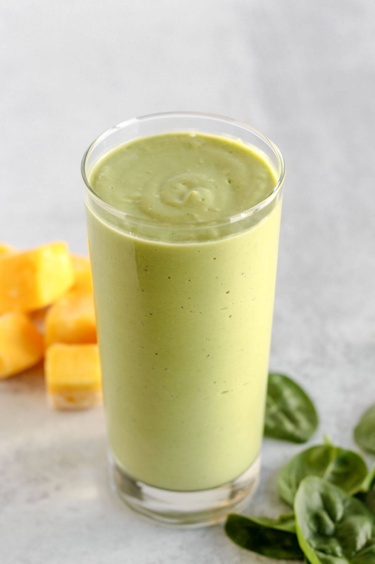 Glass with a bright green smoothie, spinach leaves and mango on the side. 