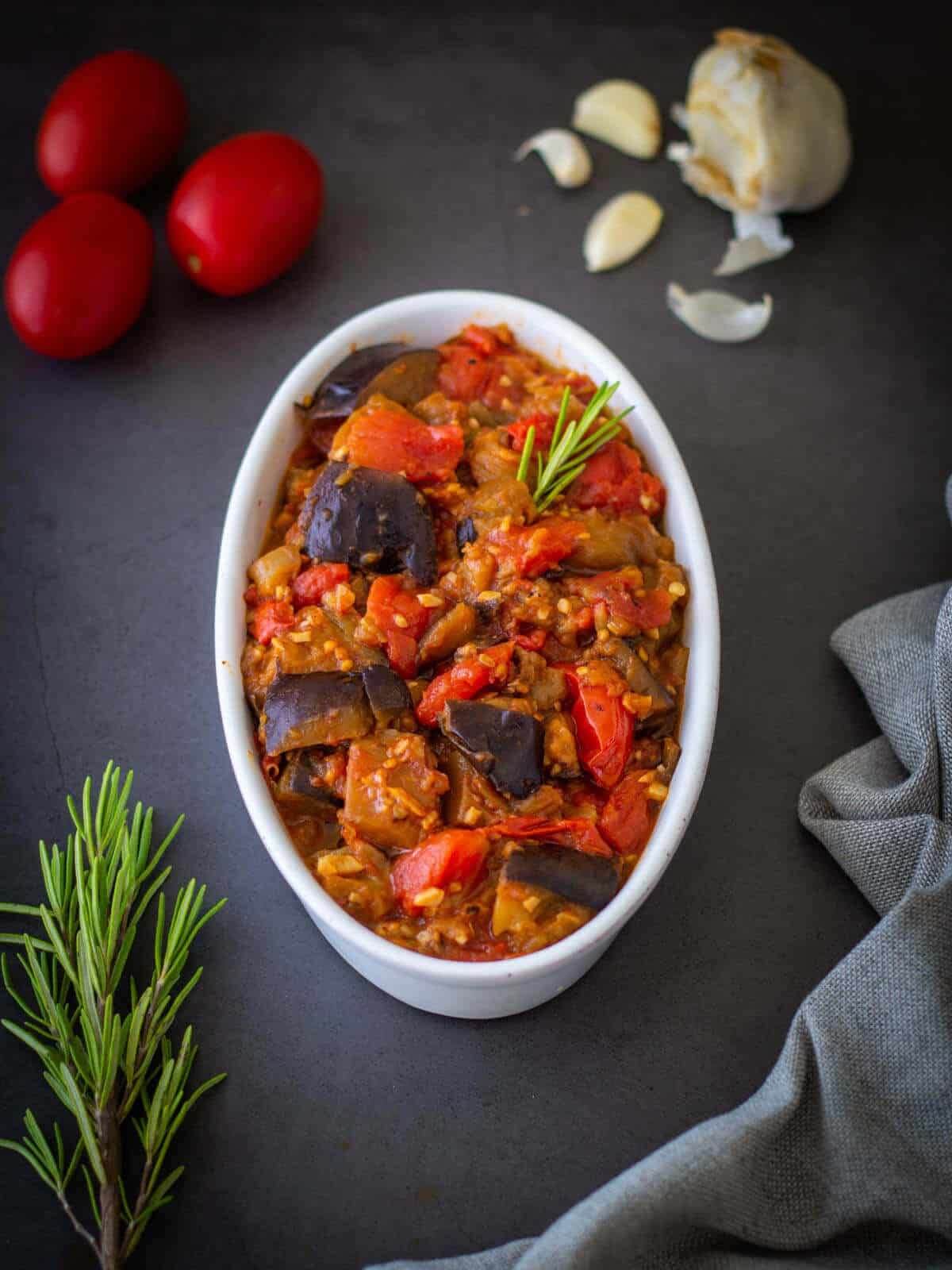 White bowl with stew. Garlic, rosemary and tomatoes on the side. 