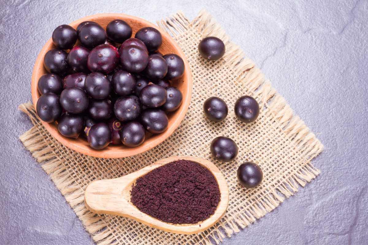 A bunch of acai berries in a wooden bowl next to a wooden spoon with dried and ground acai inside it