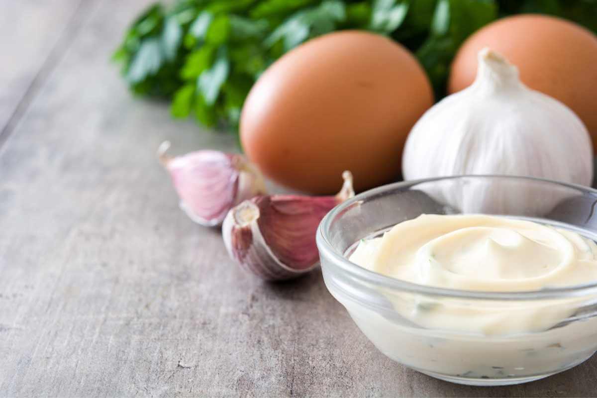 A small bowl of aioli in front of some garlic, eggs and herbs