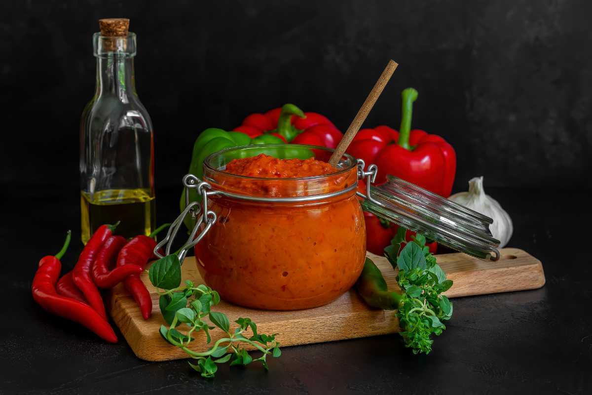 A jar of ajvar next to some bell peppers, olive oil and chili