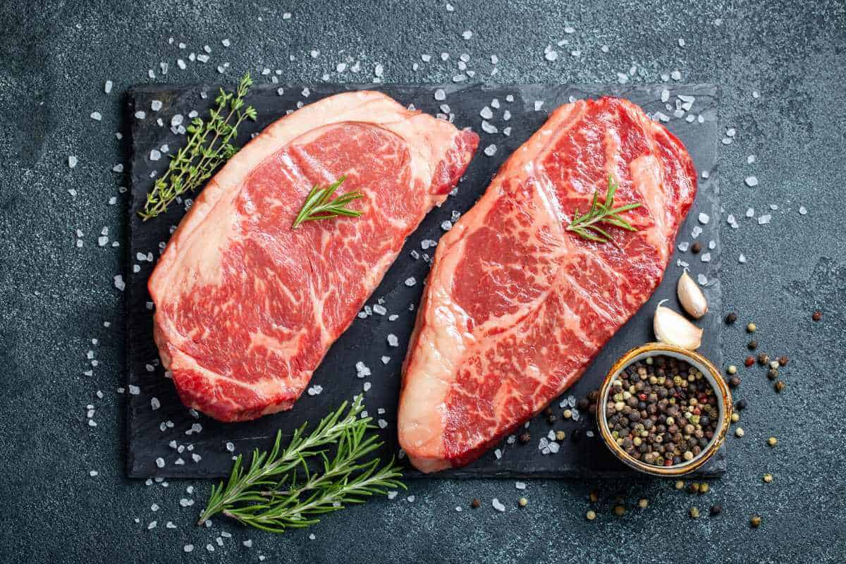 2 raw angus beef steaks on a slate board surrounded by some sea salt, black peppercorns, garlic and rosemary