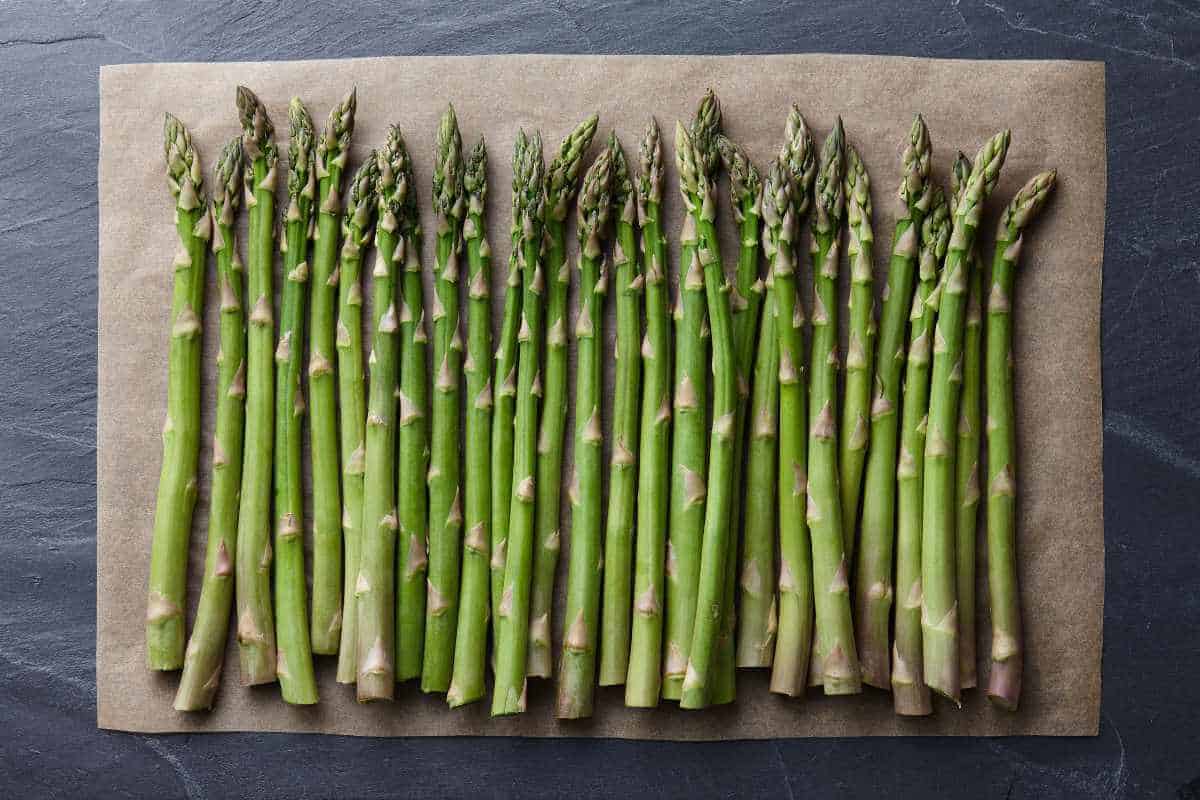 A lot of green asparagus lined up together on a baking sheet on a black slate background