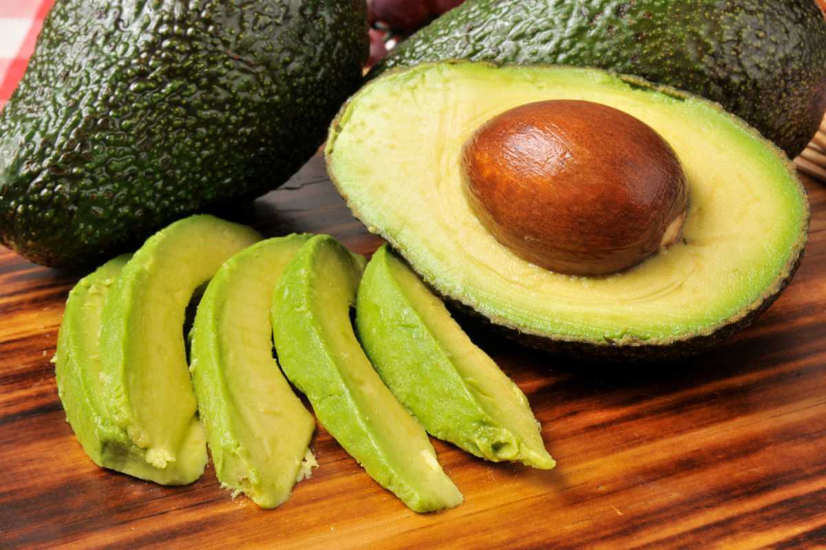 An avocado cut in half with the brown stone still inside, next to a few slices. of avocado and 2 full avocados behind