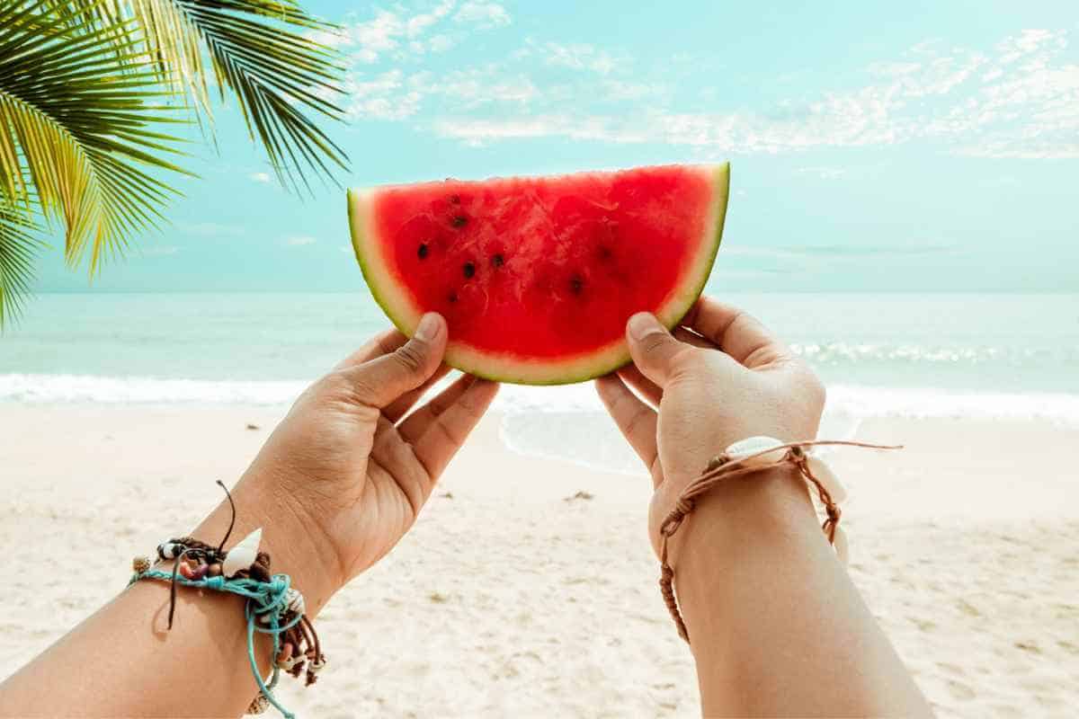 Slice of watermelon held up by two hands in front of a backdrop of the beach and sea