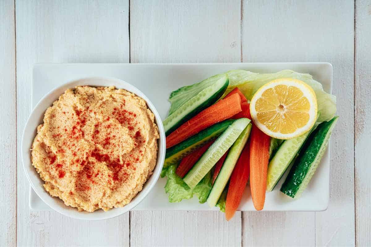 A plate of cucumber and carrot sticks with a lemon half and a bowl of hummus. A good example of beach snacks