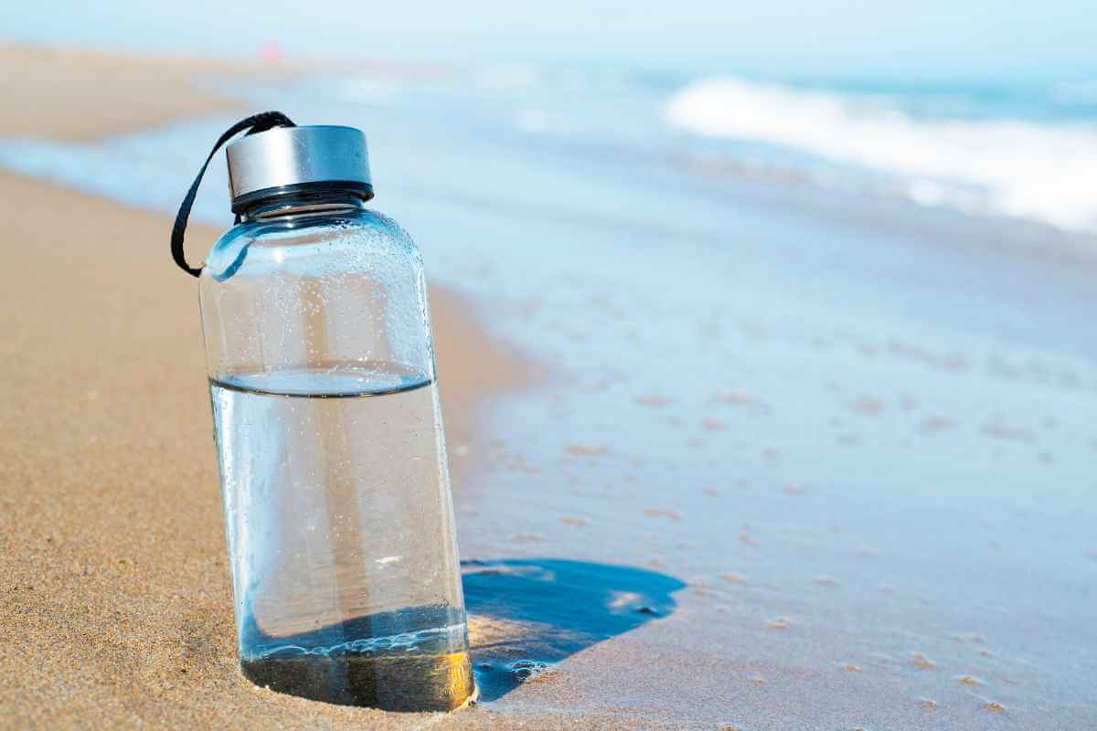 A glass bottle of water partially buried in the sand on the beach