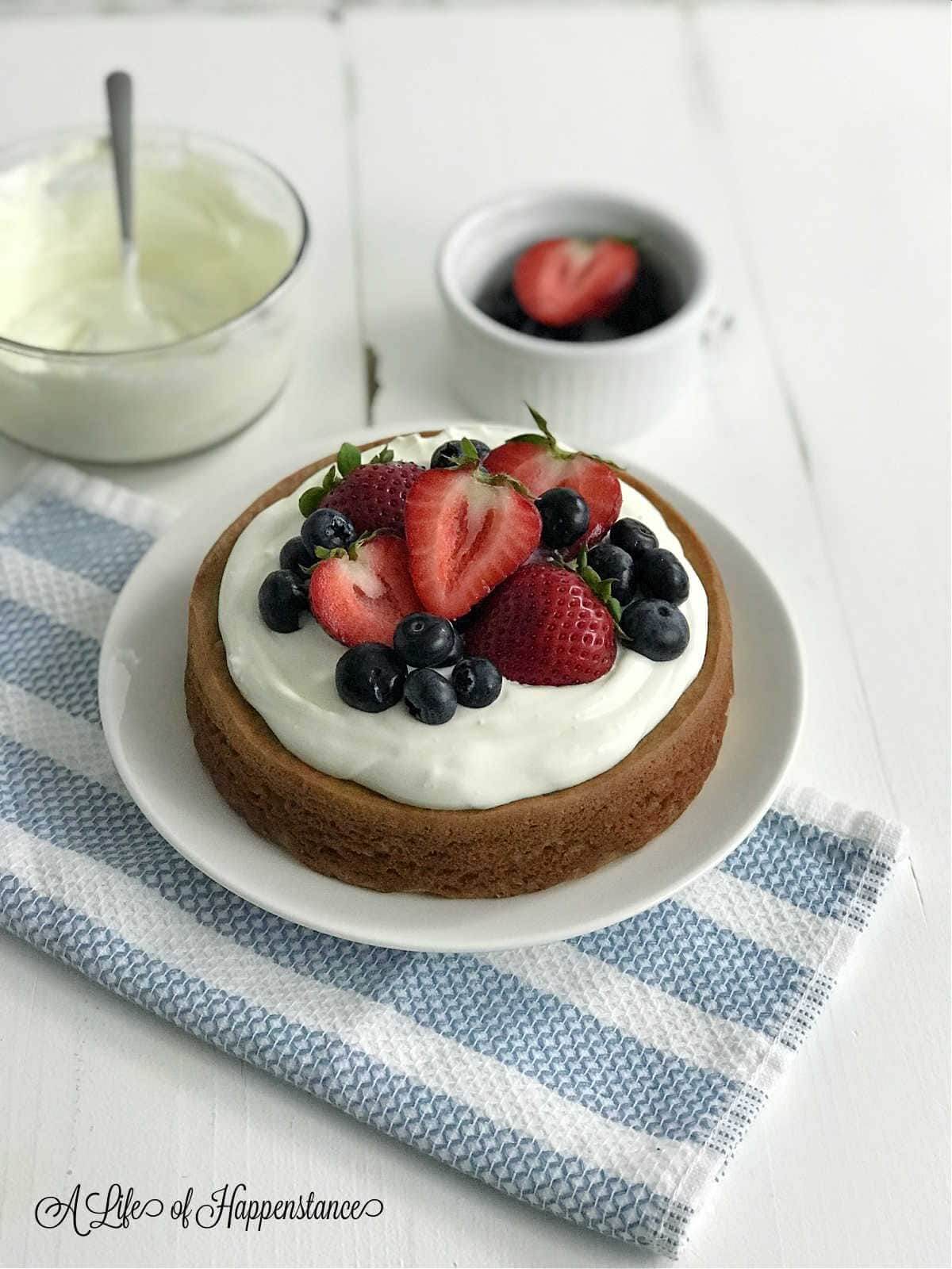 Round cake covered in whipped cream, blueberries and strawberries on a white plated. Additional whipped cream and strawberry in a small mug on the side. 