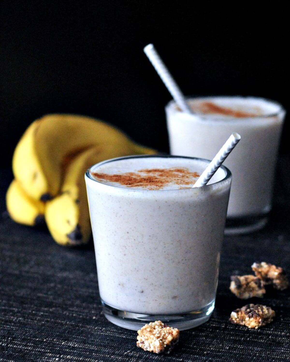 Two glasses of a white smoothie with a straw. Topped with cinnamon and some bananas on the side.