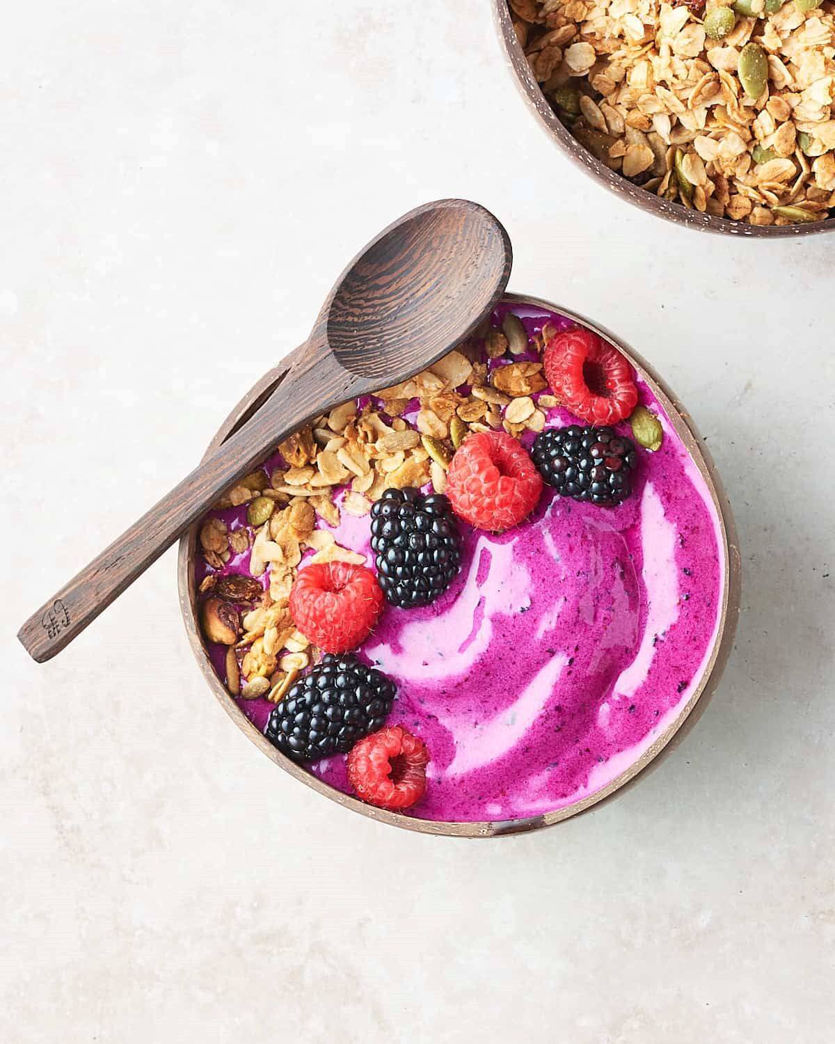 A wooden bowl with a light purple smoothie. Topped with some oats, blackberries and raspberries. 