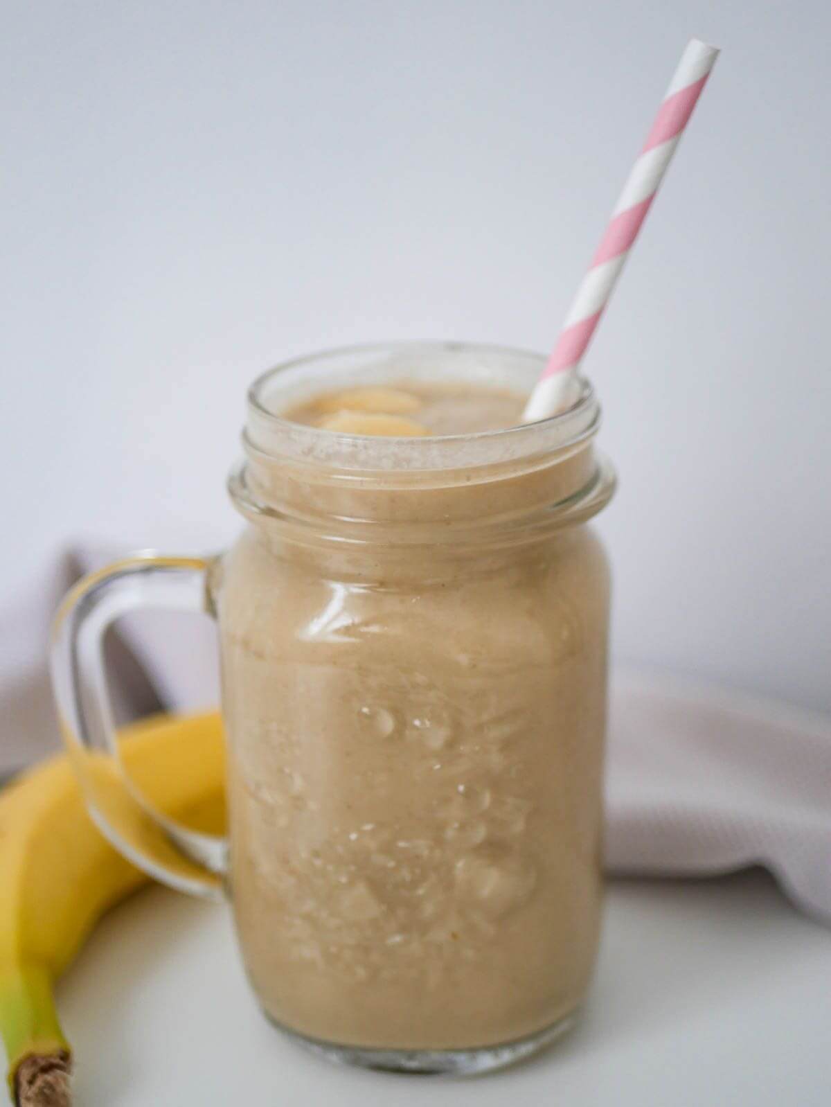 A glass with a light brown smoothie and straw. Banana on the side.