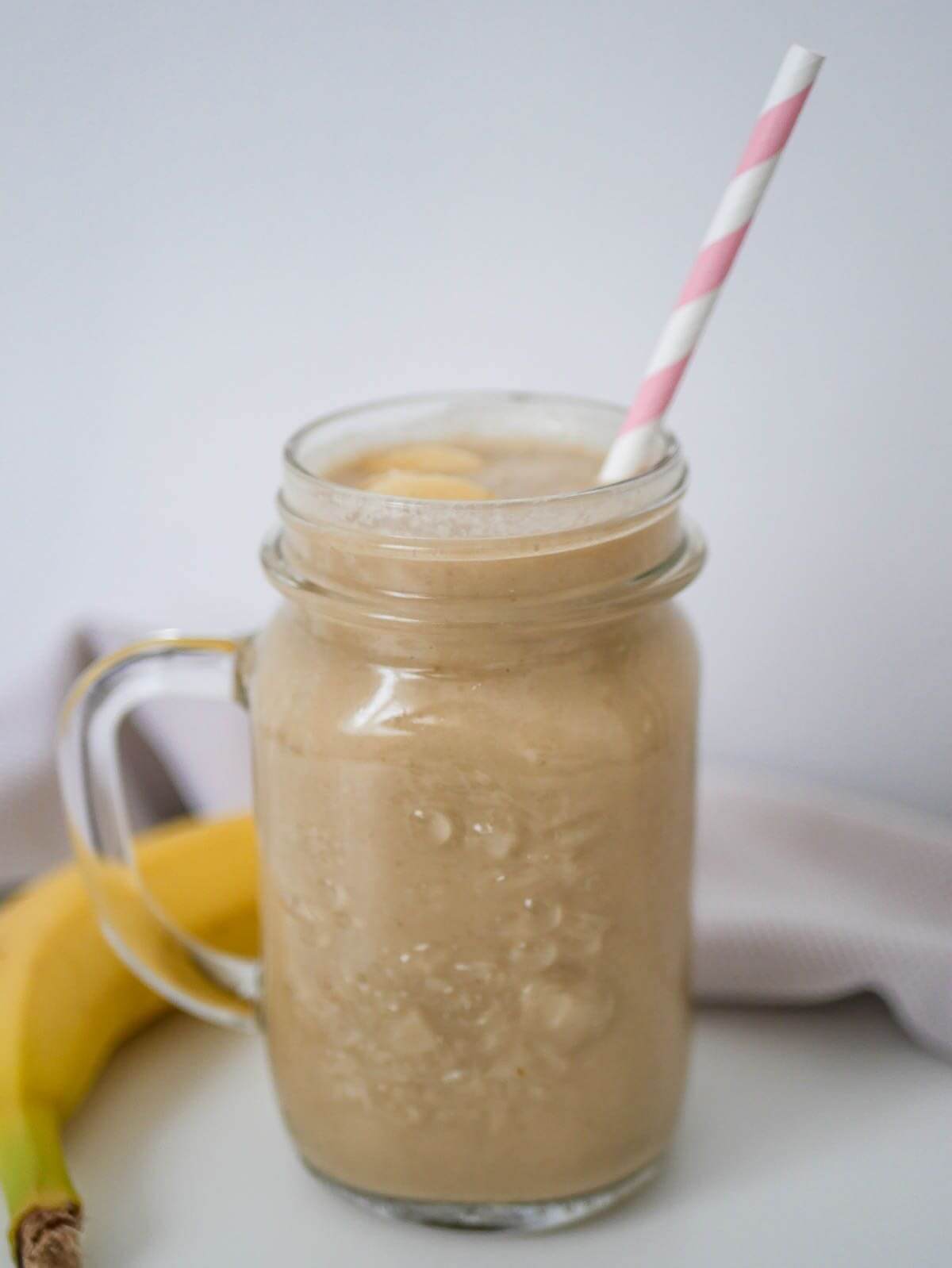 A  glass with a light brown smoothie and straw. Banana on the side. 