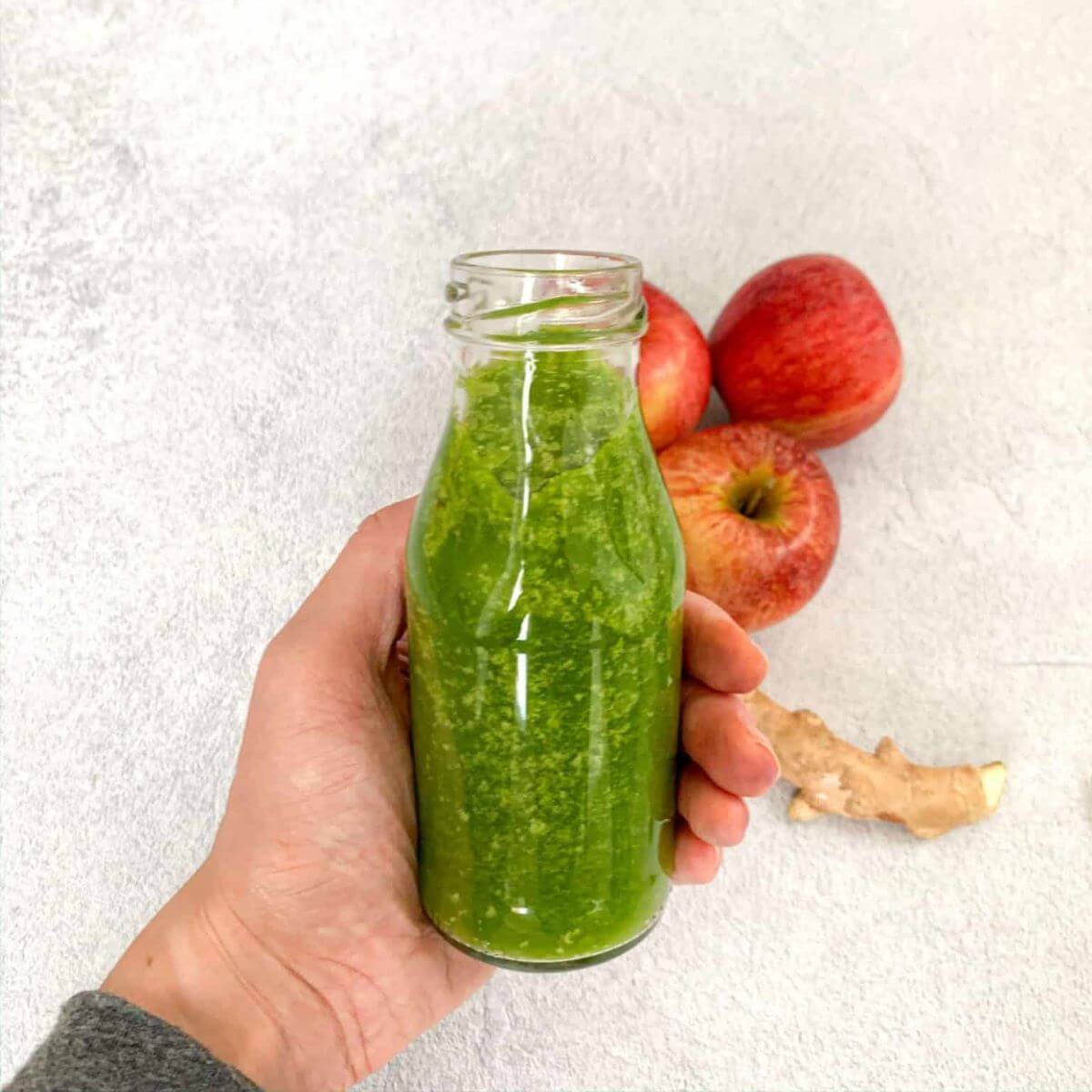 A hand holding a bottle with a green smoothie. Apples and cinnamon on the side. 