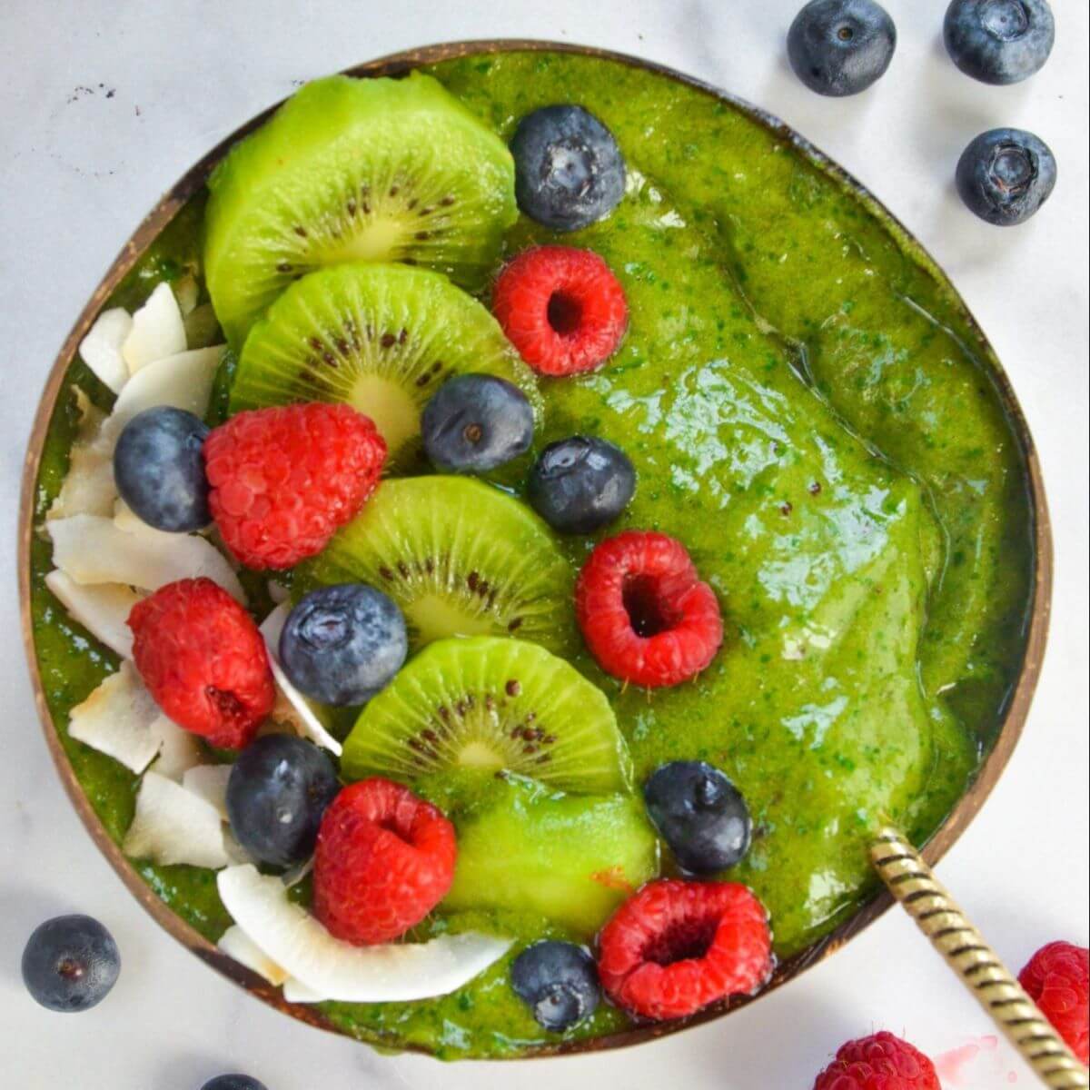 A bowl with a green smoothie topped with some sliced kiwi, raspberries, blueberries and shredded coconut.