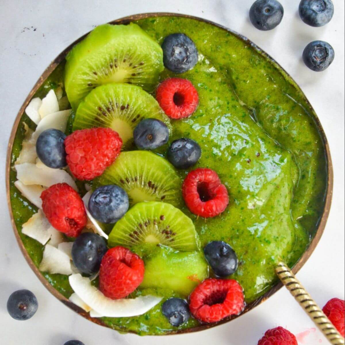 A bowl with a green smoothie topped with some sliced kiwi, raspberries, blueberries and shredded coconut. 