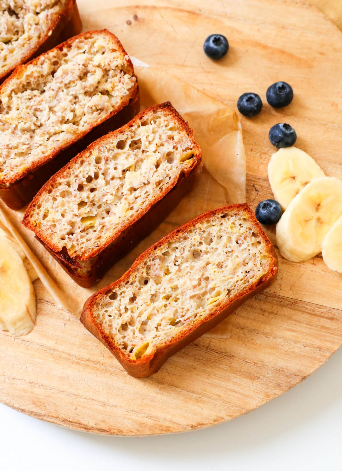 diagonal dairy free banana bread slices on a wooden board with blueberries and banana slices on the side.