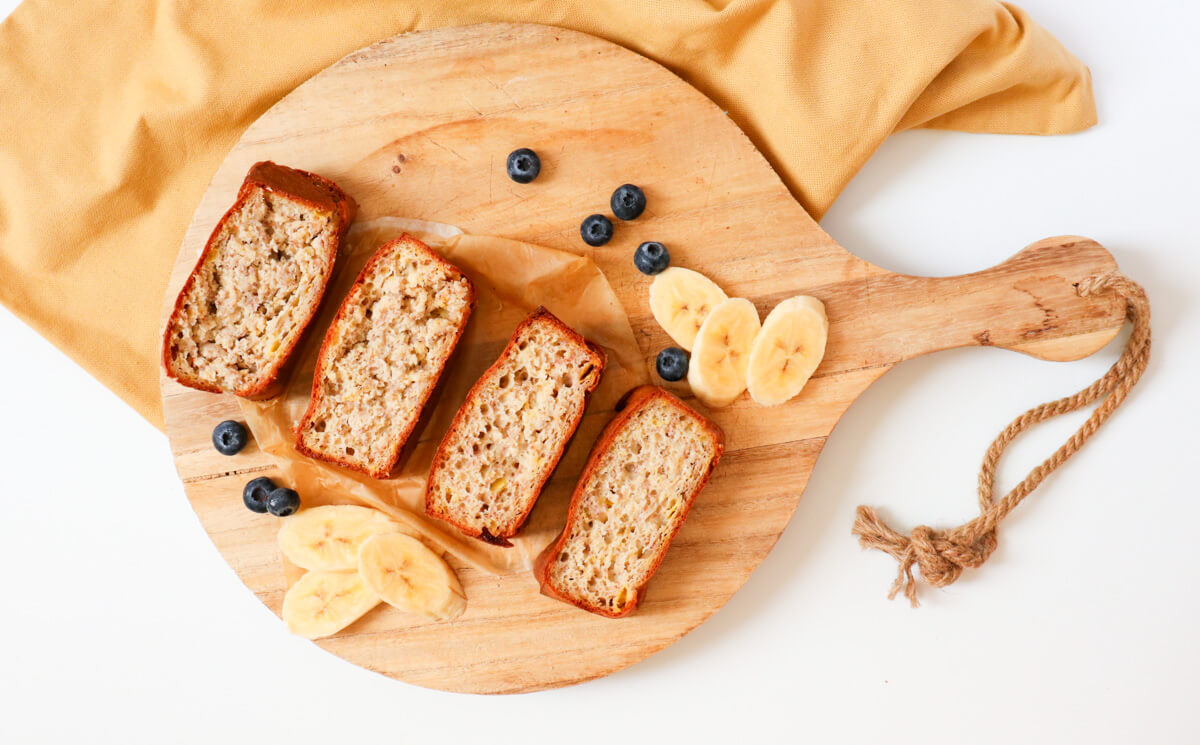 Landscape image of 4 slices of sugar free banana bread on a wooden board with banana slices and blueberries on the side.