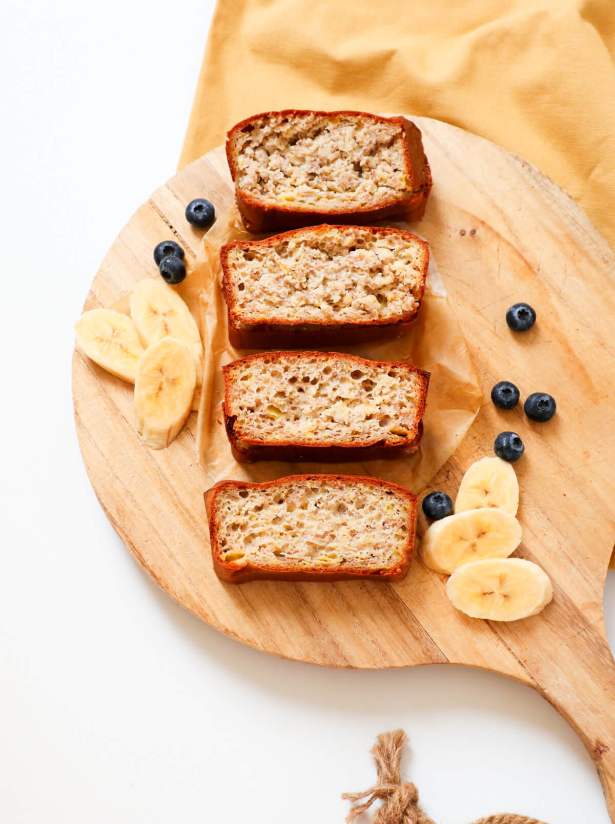 4 slices of sugar free banana bread on a wooden board with banana slices and blueberries on the side. 