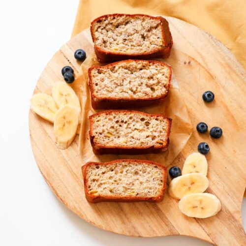 4 slices of sugar free banana bread on a wooden board with banana slices and blueberries on the side.
