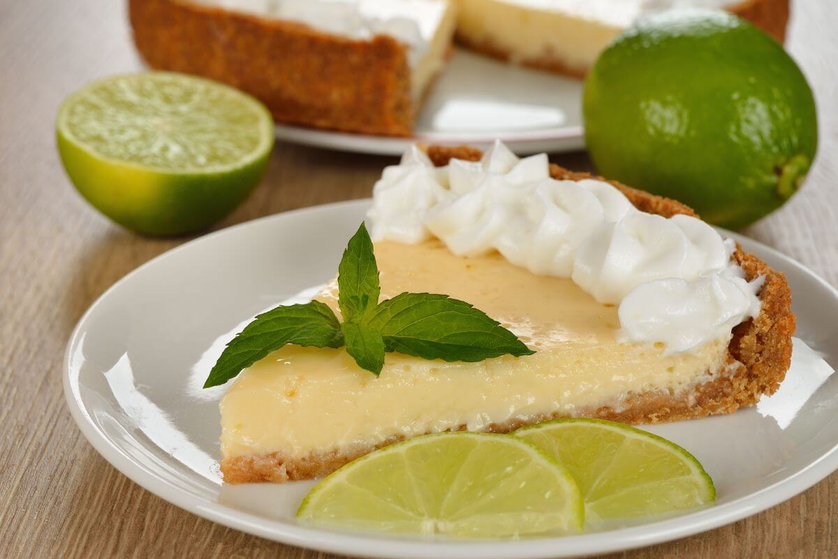 A slice of key lime pie (a delicious dessert food that stats with K) on a plate with a few limes behind and some slices of lime on the plate. The full pie is in the background