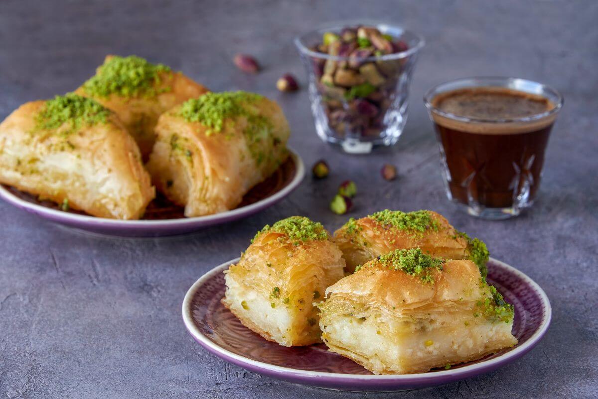 3 pieces of knafeh on a plate in front of another plate of knafeh and a glass of coffee and another glass of shelled pistachios